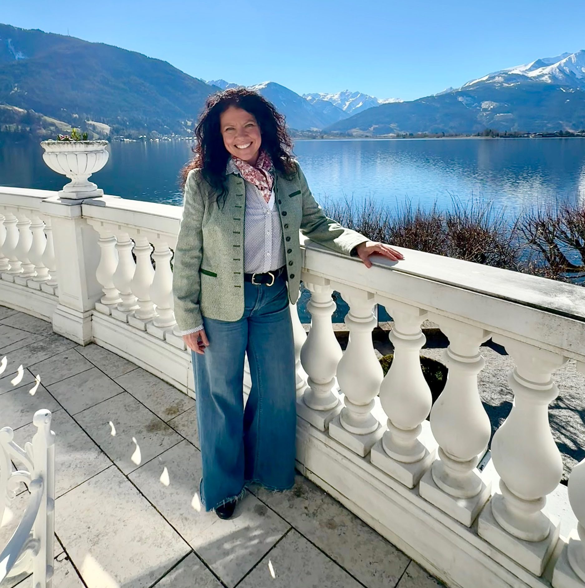 Woman standing on a terrace with white balustrades, smiling, with a lake and snow-capped mountains in the background under a clear blue sky.