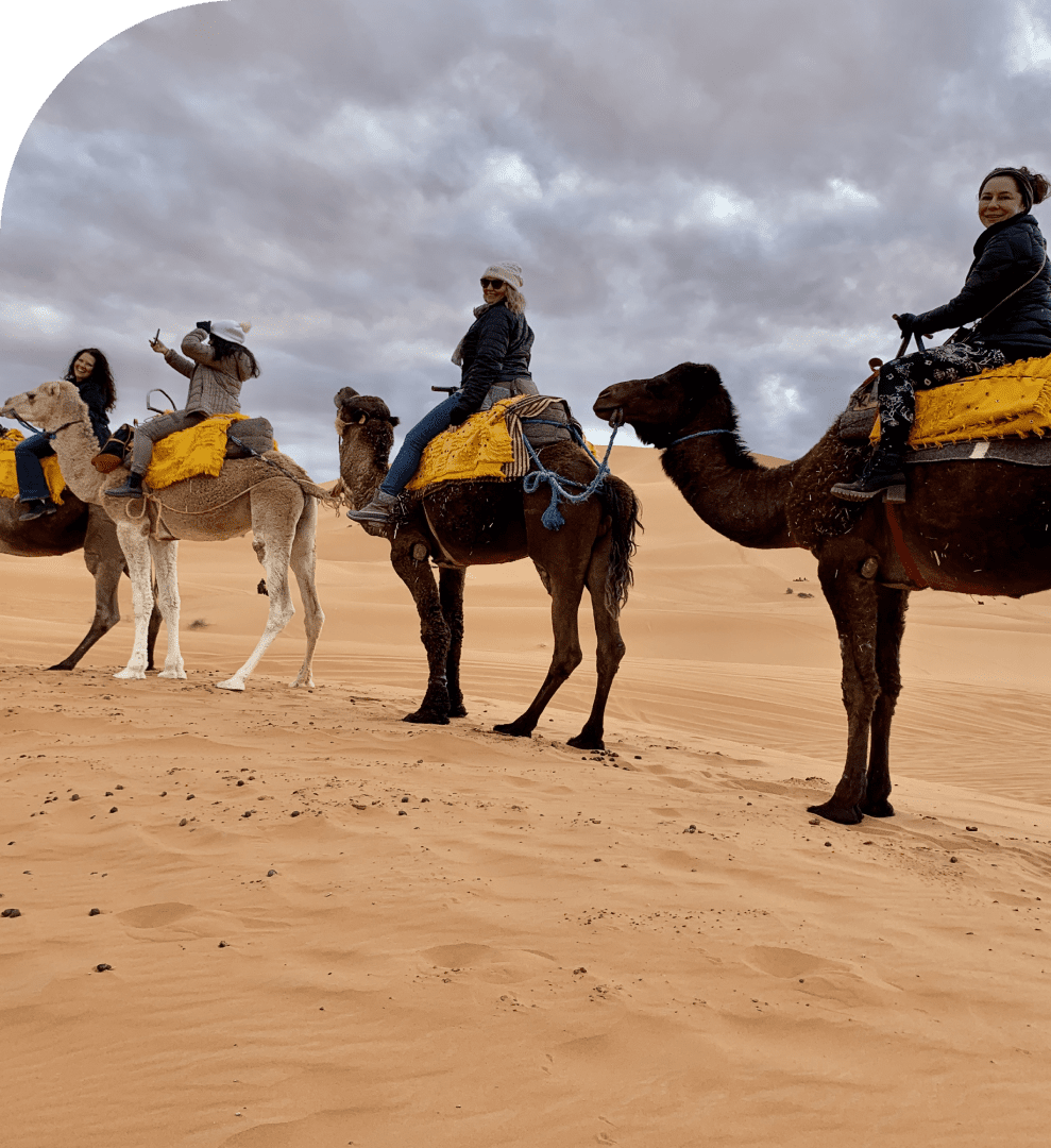 Four people ride camels with yellow blankets through a sandy desert under a cloudy sky.