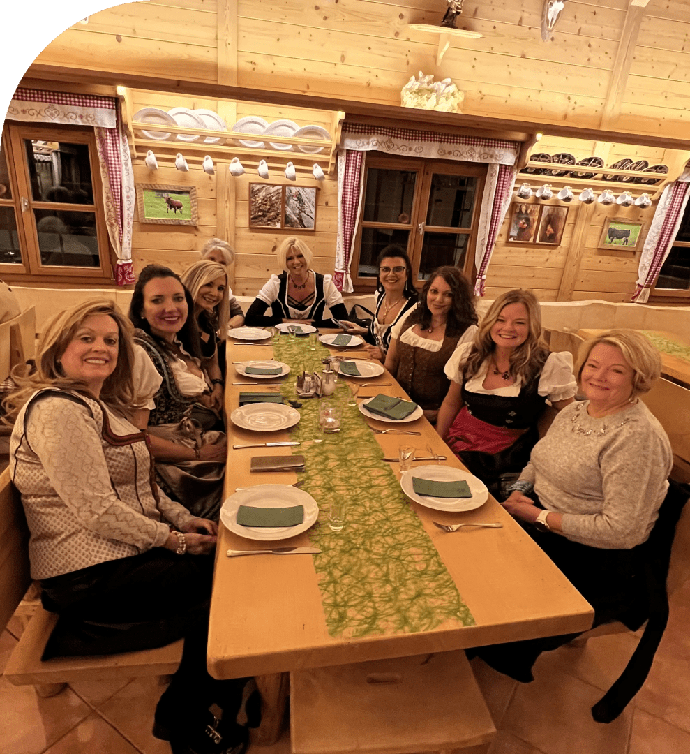 2 Eight women are seated around a wooden dining table in a cozy, rustic room with wood-paneled walls, ready for a meal.