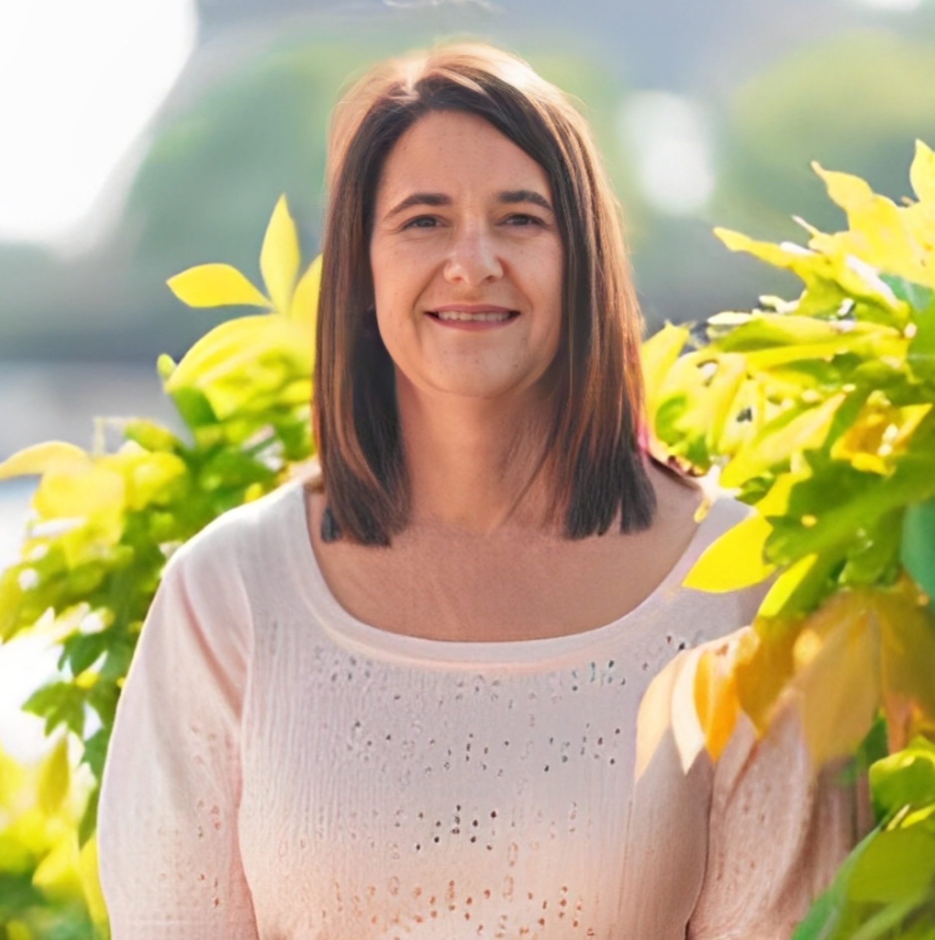 A woman with straight brown hair, wearing a light pink top, stands outdoors in front of green and yellow foliage, smiling at the camera.
