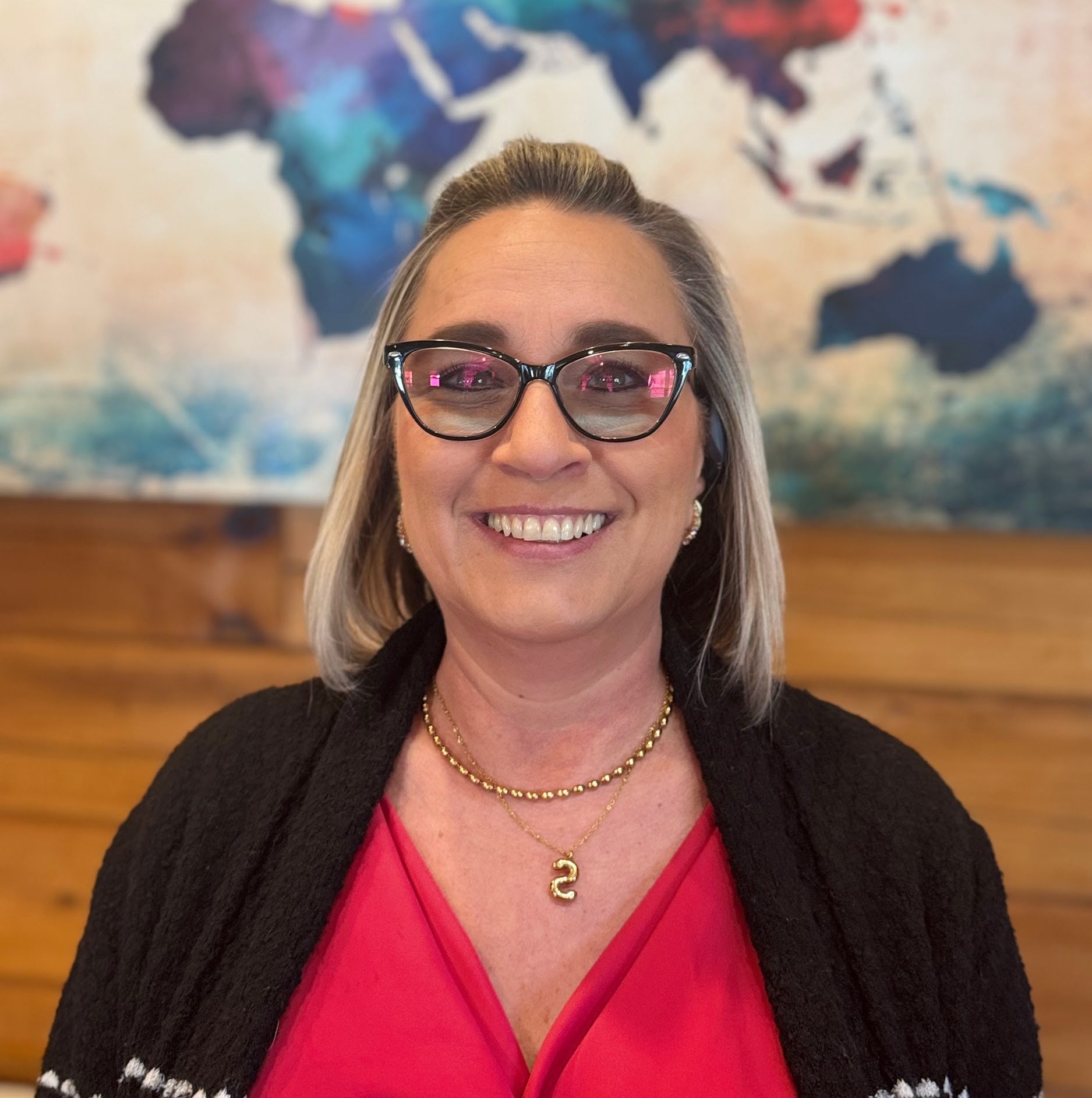 A woman with shoulder-length blonde hair and glasses smiles at the camera; she is wearing a red top, black sweater, gold necklace, and stands in front of a world map.