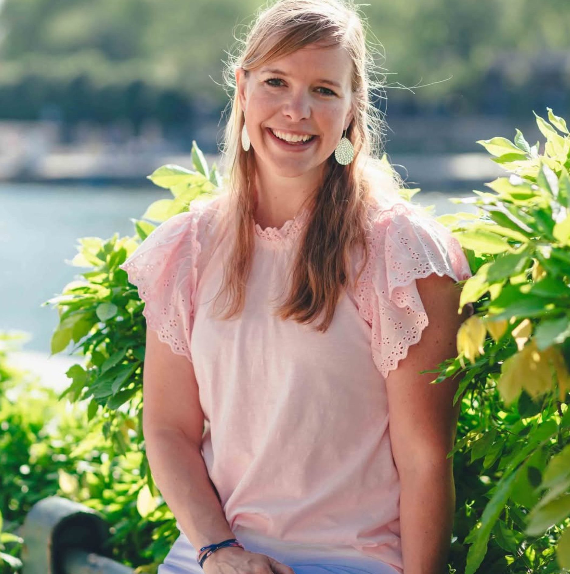 Woman with long blonde hair in a light pink short-sleeve top sits outdoors on a bench, surrounded by green plants, smiling at the camera.