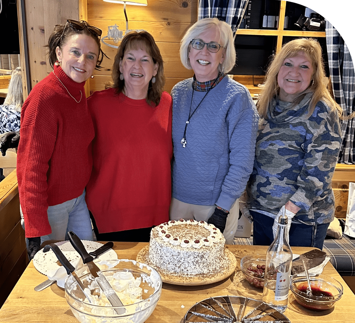 Four women stand together smiling behind a table with a decorated cake, dessert plates, utensils, and a bottle in a cozy, wood-paneled room.