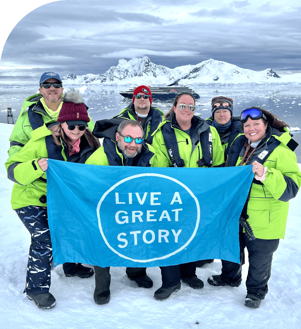 Seven people in green jackets stand on snow holding a blue "Live A Great Story" flag, with snowy mountains and water in the background.