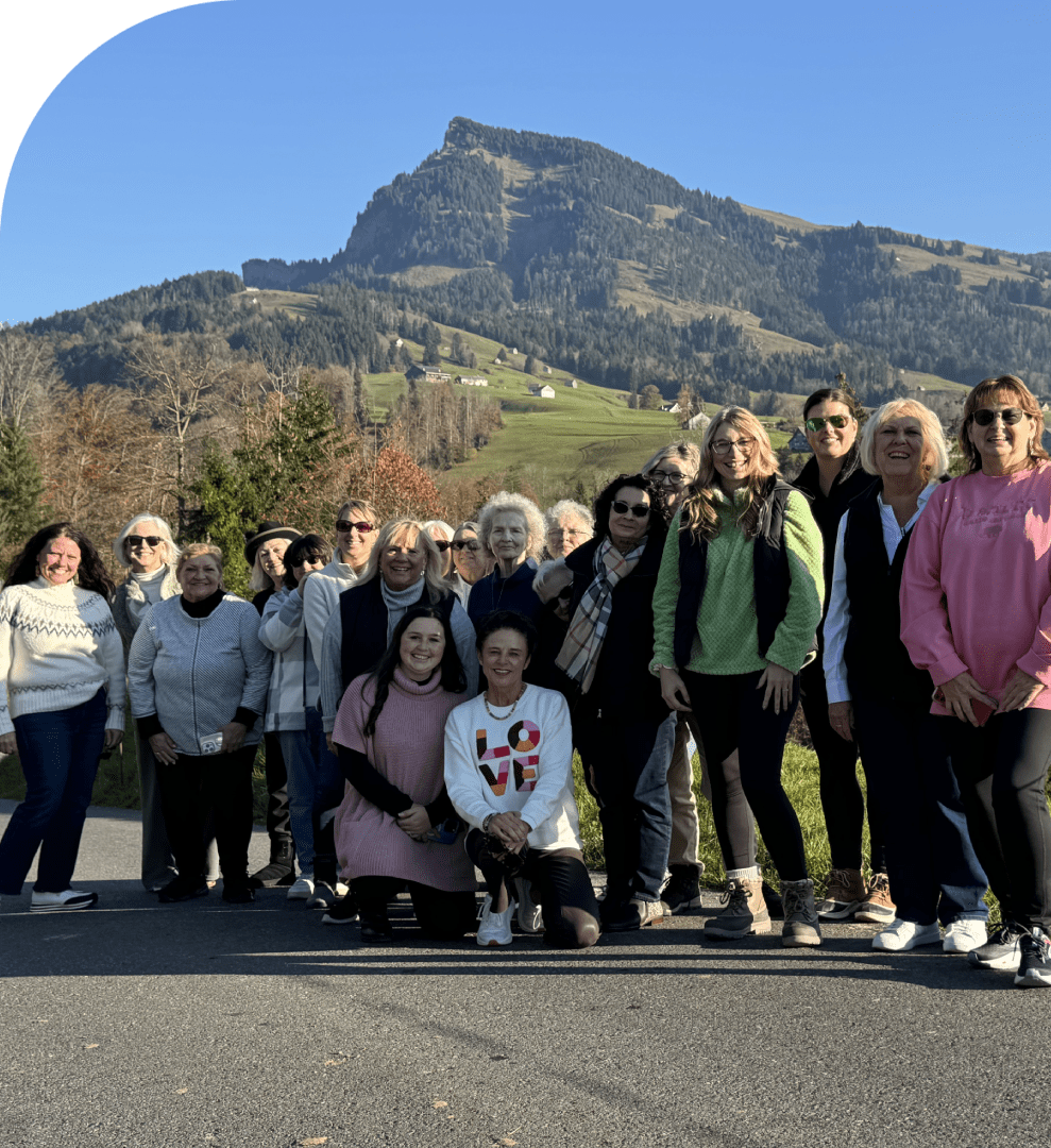 Frame-1618873625-2 A group of people poses for a photo outdoors on a sunny day with a mountain and green hillside in the background.