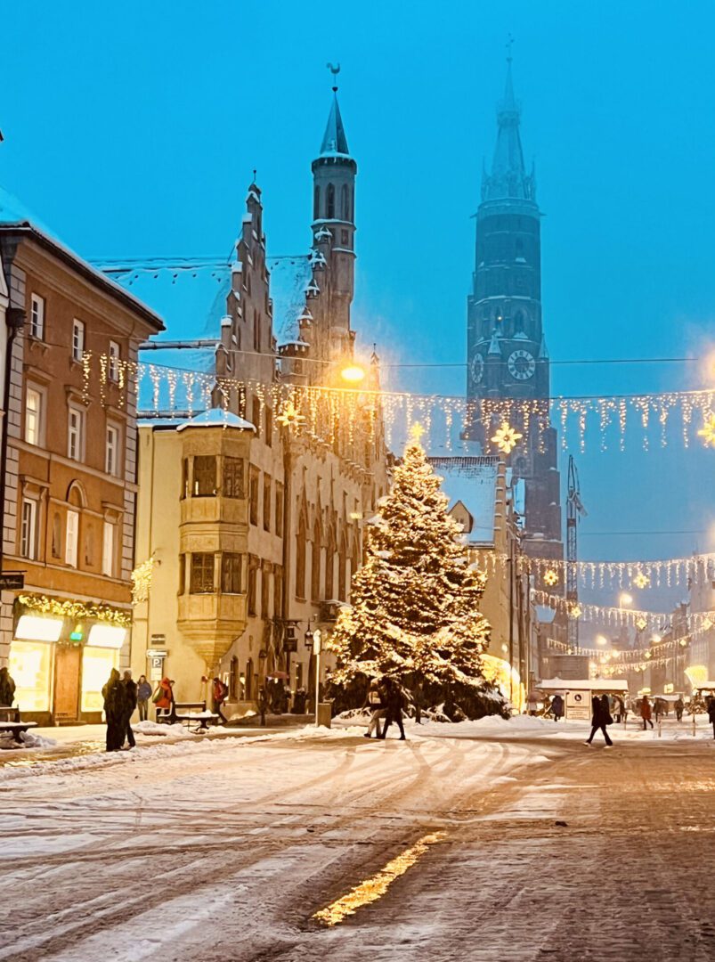 A snow-covered town square features a large decorated Christmas tree, historic buildings, illuminated shops, and string lights overhead during winter.