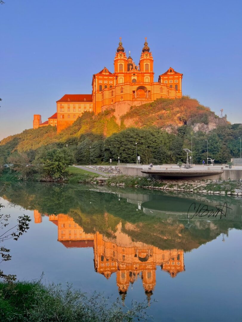 A baroque-style abbey on a hill is reflected in the calm river below, surrounded by green trees under a clear blue sky at sunset.